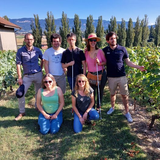 Posant fièrement devant les vignes du Domaine des Bonnettes, à Lully, dans la campagne genevoise, l'ensemble des participantes et participants voyants, malvoyants et aveugles de la sortie du 12 septembre 2021 des Jeunes FSA Genève. De gauche à droite: Etienne Rallu, Alexandre Turc, Alex Bernier (sa canne blanche dépliée à la main), Céline Witschard (sa canne blanche dépliée à la main), Thibault Trancart et, à genoux ou accroupie dans l'herbe, Hélène Reymermier et Francesca Maigre.Toute l'équipe est souriante, debout dans l'herbe devant les vignes sous un soleil radieux. Au second plan, derrière eux, légèrement en contrebas, la cime d'une allée de cyprès et, au fond au loin, la silhouette montagneuse du Salève qui se dresse sous un ciel bleu azur.